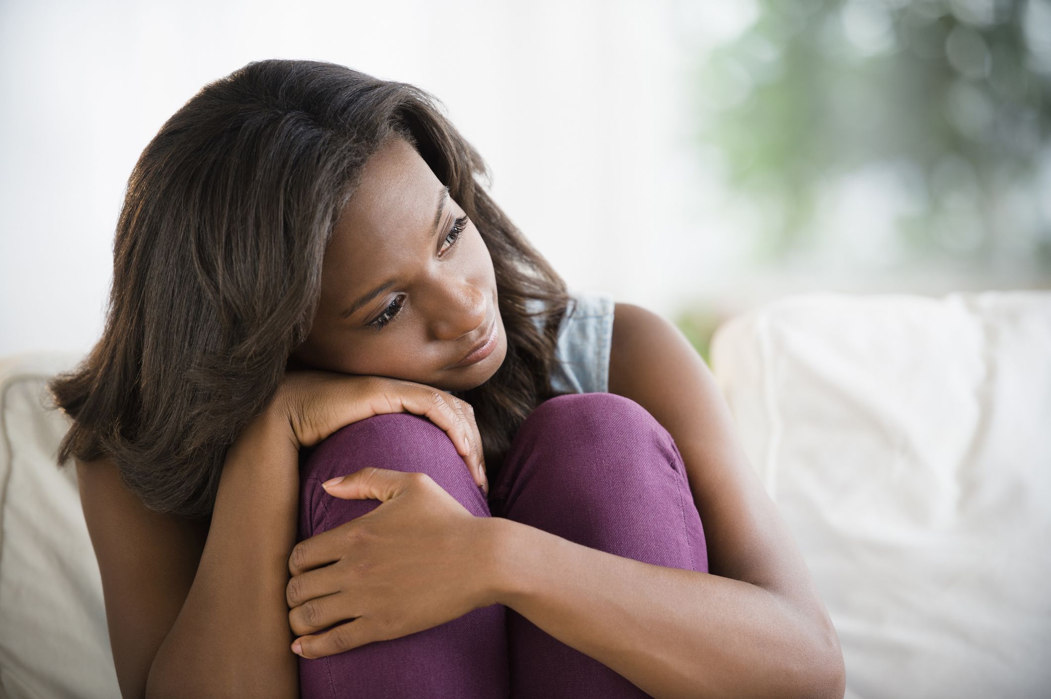 Woman seated on a sofa with her head resting on her folded arms and knees.