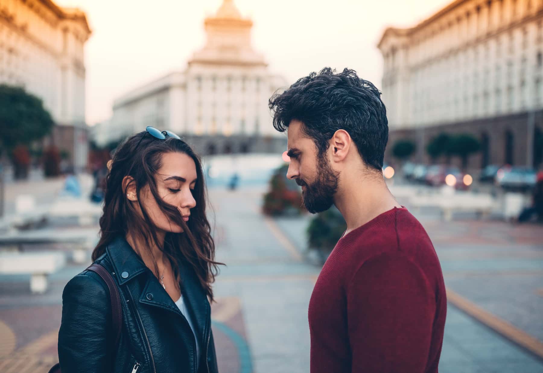 Two adults facing each other in a city square, with the woman looking down.